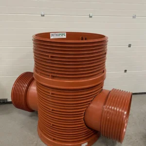 Stor orange plaströrsarmatur med tre öppningar och räfflade sidor, märkt "INFRAPIPE", stående upprätt på ett betonggolv framför en beige garagedörr.