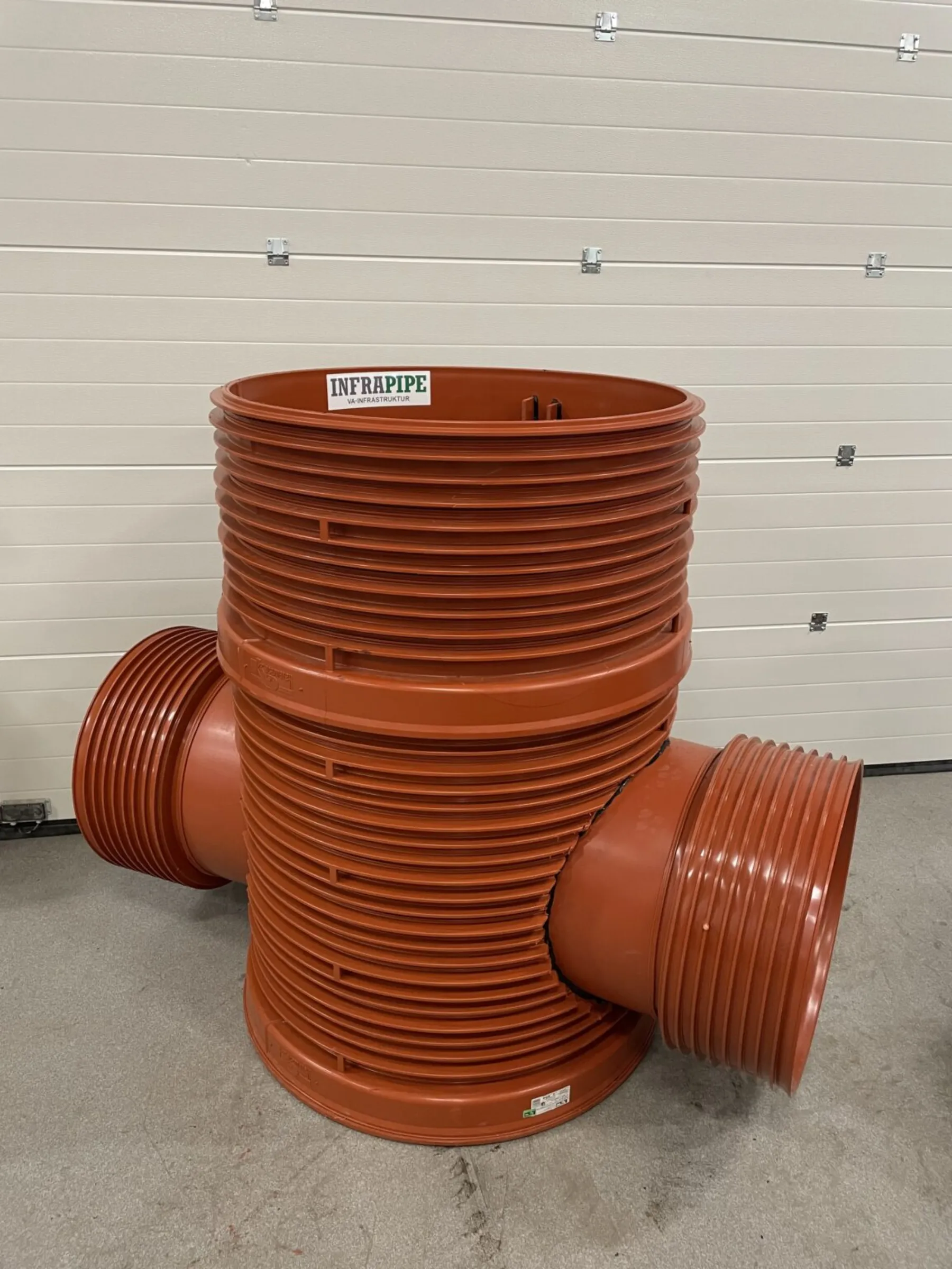 Stor orange plaströrsarmatur med tre öppningar och räfflade sidor, märkt "INFRAPIPE", stående upprätt på ett betonggolv framför en beige garagedörr.