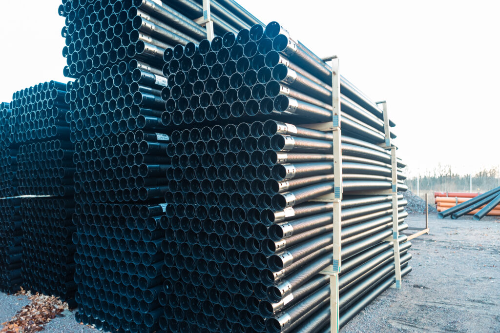 Stora staplar av svarta PP markrör 110 mm x 6 meter är prydligt ordnade och säkrade med metallramar på en grusyta utomhus, med några orange rör synliga i bakgrunden.
