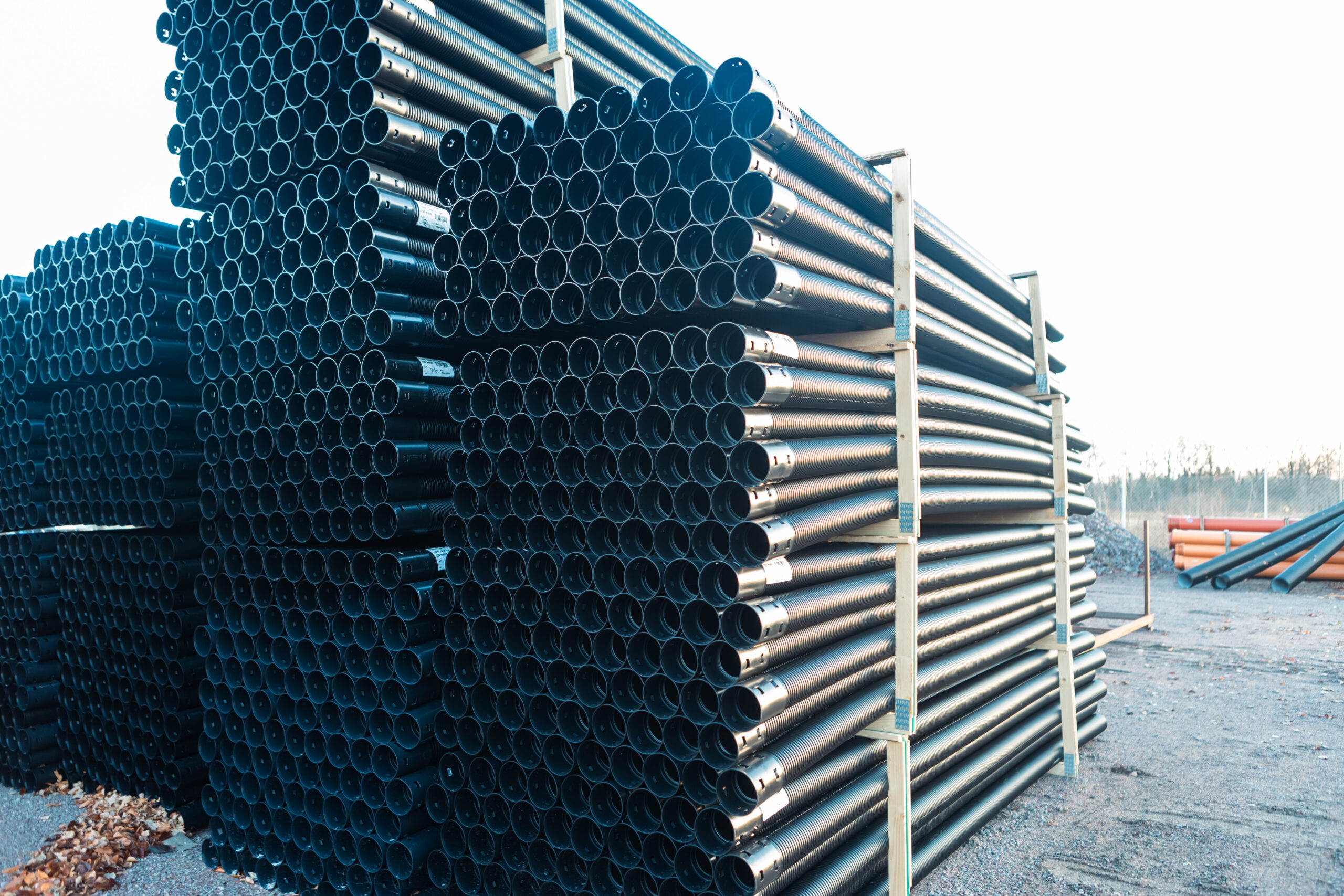 Stora staplar av svarta PP markrör 110 mm x 6 meter är prydligt ordnade och säkrade med metallramar på en grusyta utomhus, med några orange rör synliga i bakgrunden.