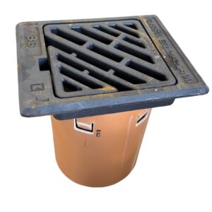 Ett stormavloppsgaller av metall med diagonala lameller är fäst vid ett runt brunt dräneringsrör, isolerat på en vit bakgrund.