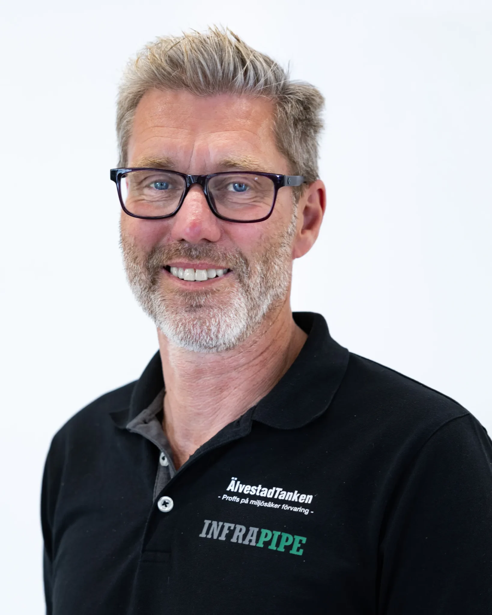 En medelålders man med grått hår och skägg, bär glasögon med svart båge och en svart polotröja med ÄlvestadTankens och INFRAPIPE:s logotyper, ler mot en vit bakgrund.