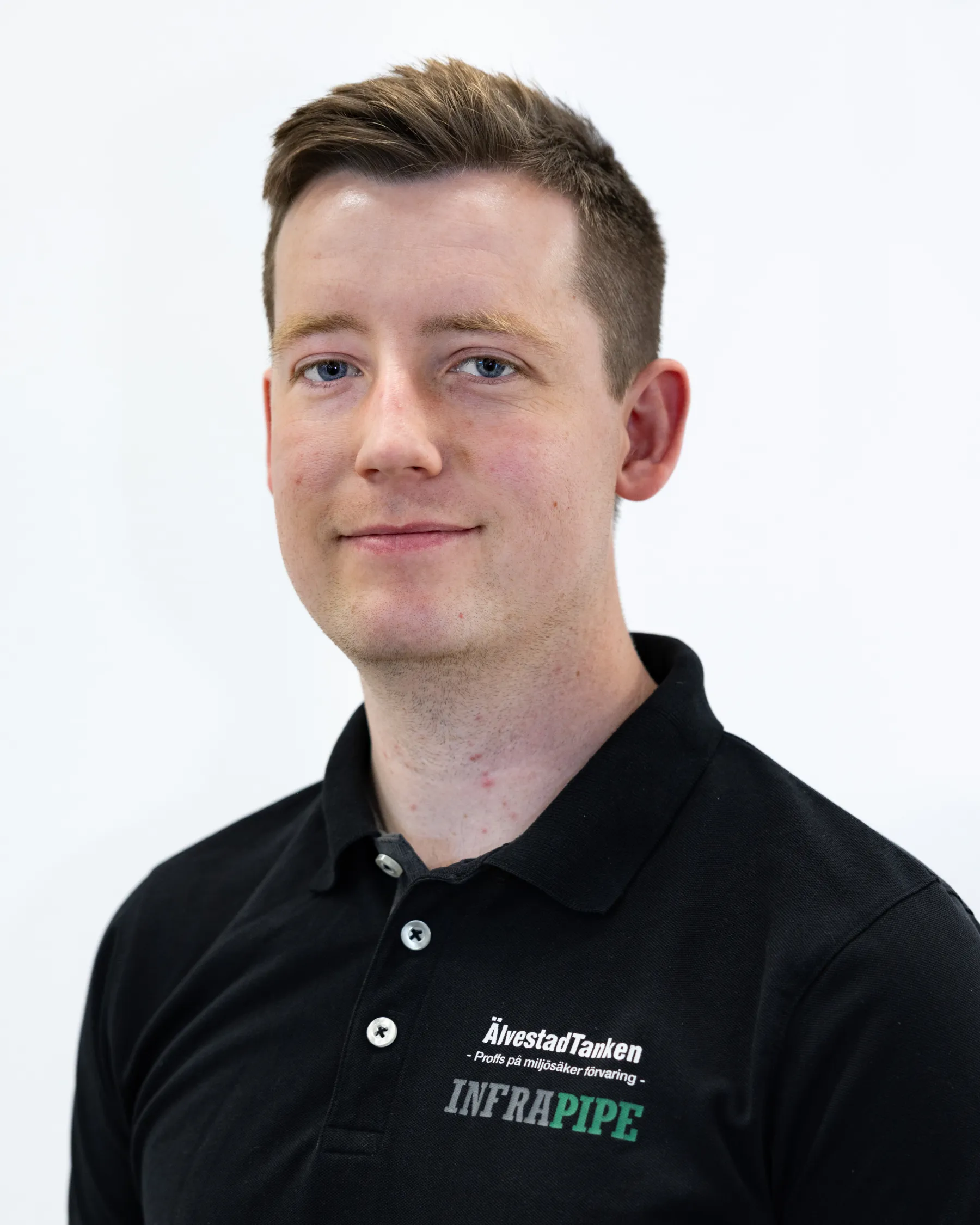 En ung man med kort brunt hår bär en svart polotröja med ÅlvestadTankern INFRAPIPE-logotyper. Han står mot en vit bakgrund och ler lite mot kameran.