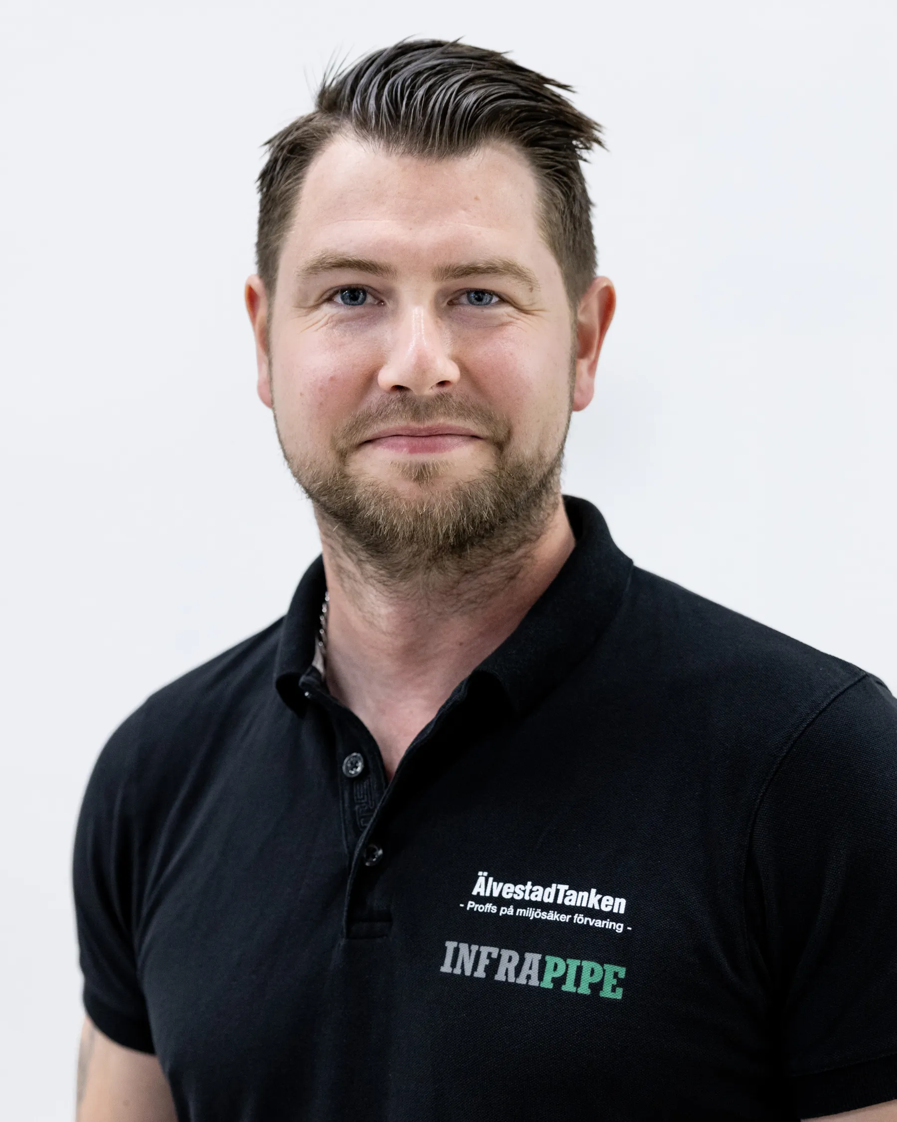 En man med kort brunt hår och vältrimmat skägg, klädd i svart pikétröja med ÄlvestadTanken och INFRAPIPE-logotyper, står framför en vit bakgrund och ler lätt.