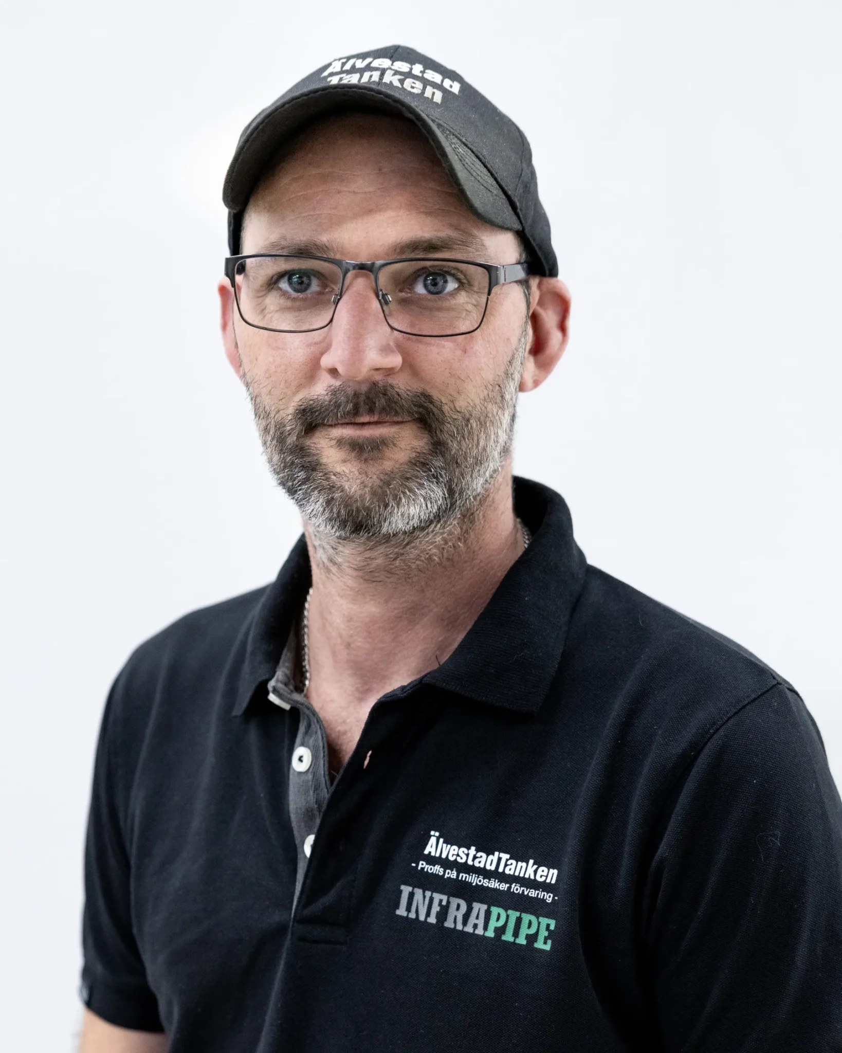 En man med skägg och glasögon, iklädd svart keps och svart polotröja med logotyperna ÄlvestadTanken och INFRAPIPE, står mot en vit bakgrund.