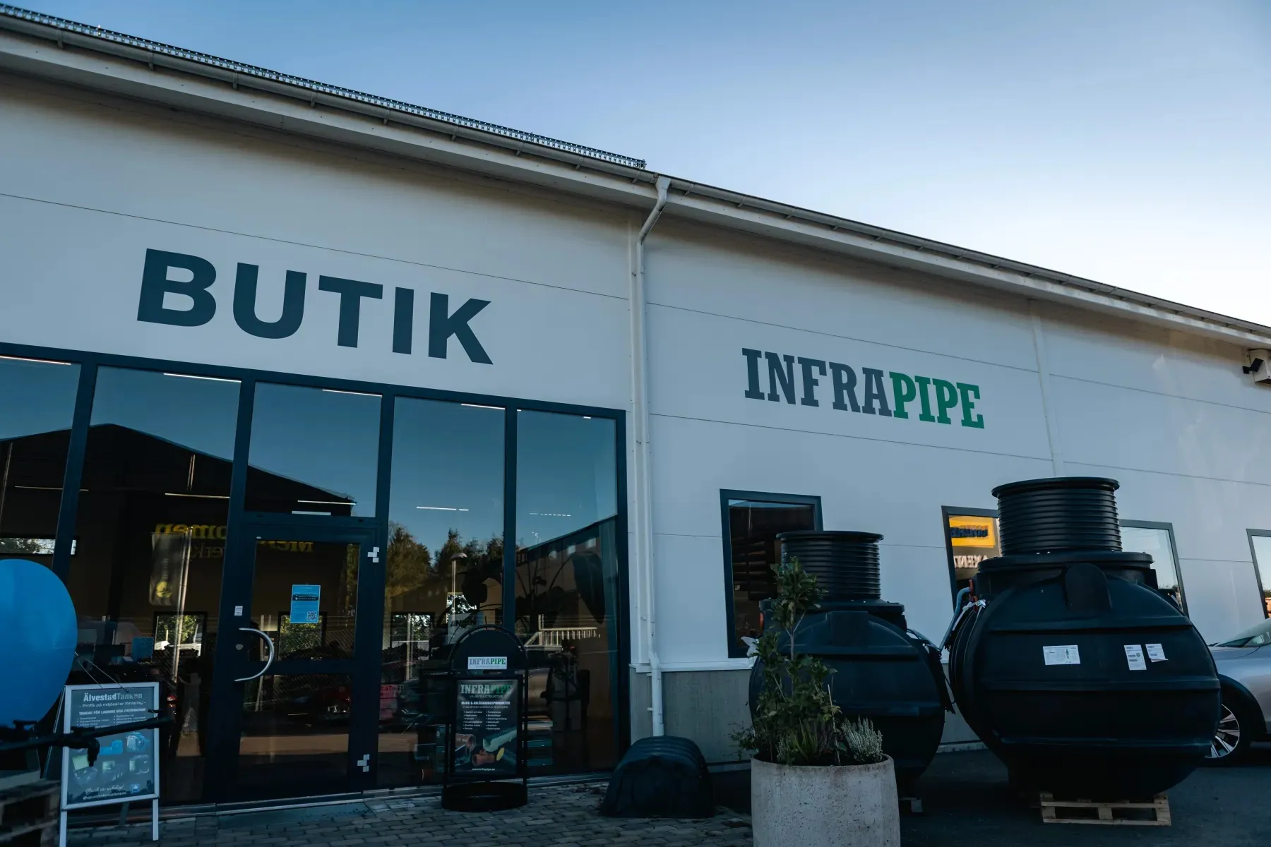 Vy från utsidan av en butik med skyltarna BUTIK och INFRAPIPE. Stora svarta industrirör eller tankar visas utomhus bredvid byggnaden under en klar himmel.
