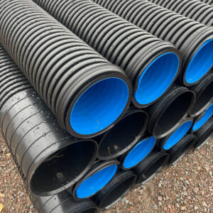 Rader av stora svarta plaströr med ljusblå innerfoder, identifierade som Evosan vägtrumma 216/250mm - 6 meter, är staplade på en grusyta och sannolikt avsedda för dränering eller byggnadsändamål.