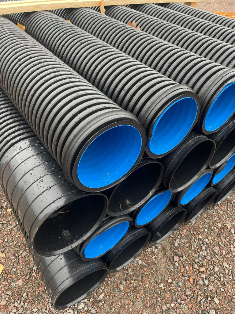 Rader av stora svarta plaströr med ljusblå innerfoder, identifierade som Evosan vägtrumma 216/250mm - 6 meter, är staplade på en grusyta och sannolikt avsedda för dränering eller byggnadsändamål.