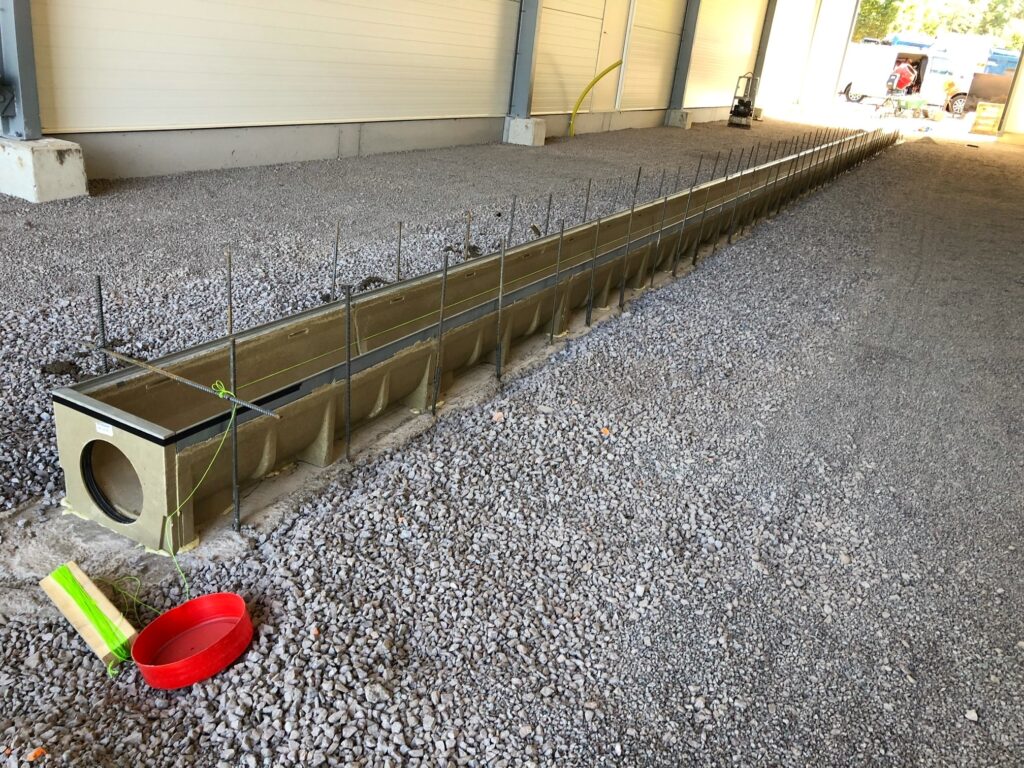 En Sandfång BASE 300 mm 0,5 m dräneringsränna med metallarmering är installerad på grus i en delvis uppförd byggnad. I närheten finns en röd skål, ett grönvitt block och byggmaterial i bakgrunden.
