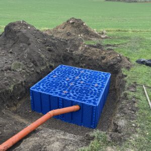 En stor blå Fördröjningsmagasin 1600 liter är delvis nedgrävd i ett rektangulärt hål, ansluten till ett orange rör. Området runtomkring har gräs, jordhögar och öppna fält i bakgrunden.