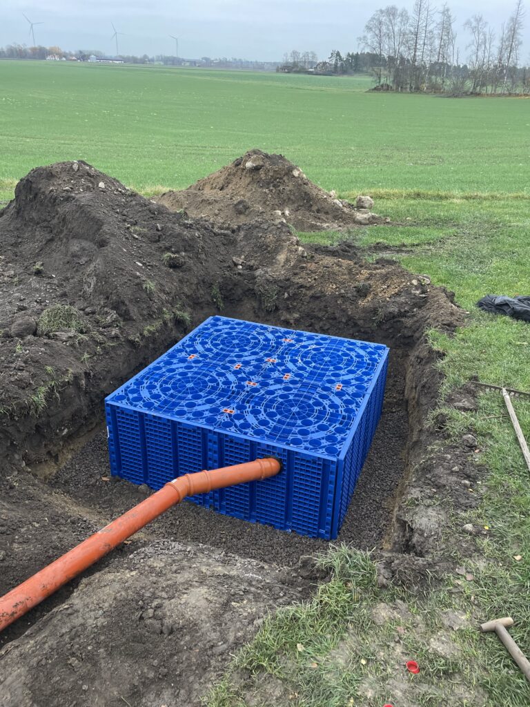 En stor blå Fördröjningsmagasin 1600 liter är delvis nedgrävd i ett rektangulärt hål, ansluten till ett orange rör. Området runtomkring har gräs, jordhögar och öppna fält i bakgrunden.