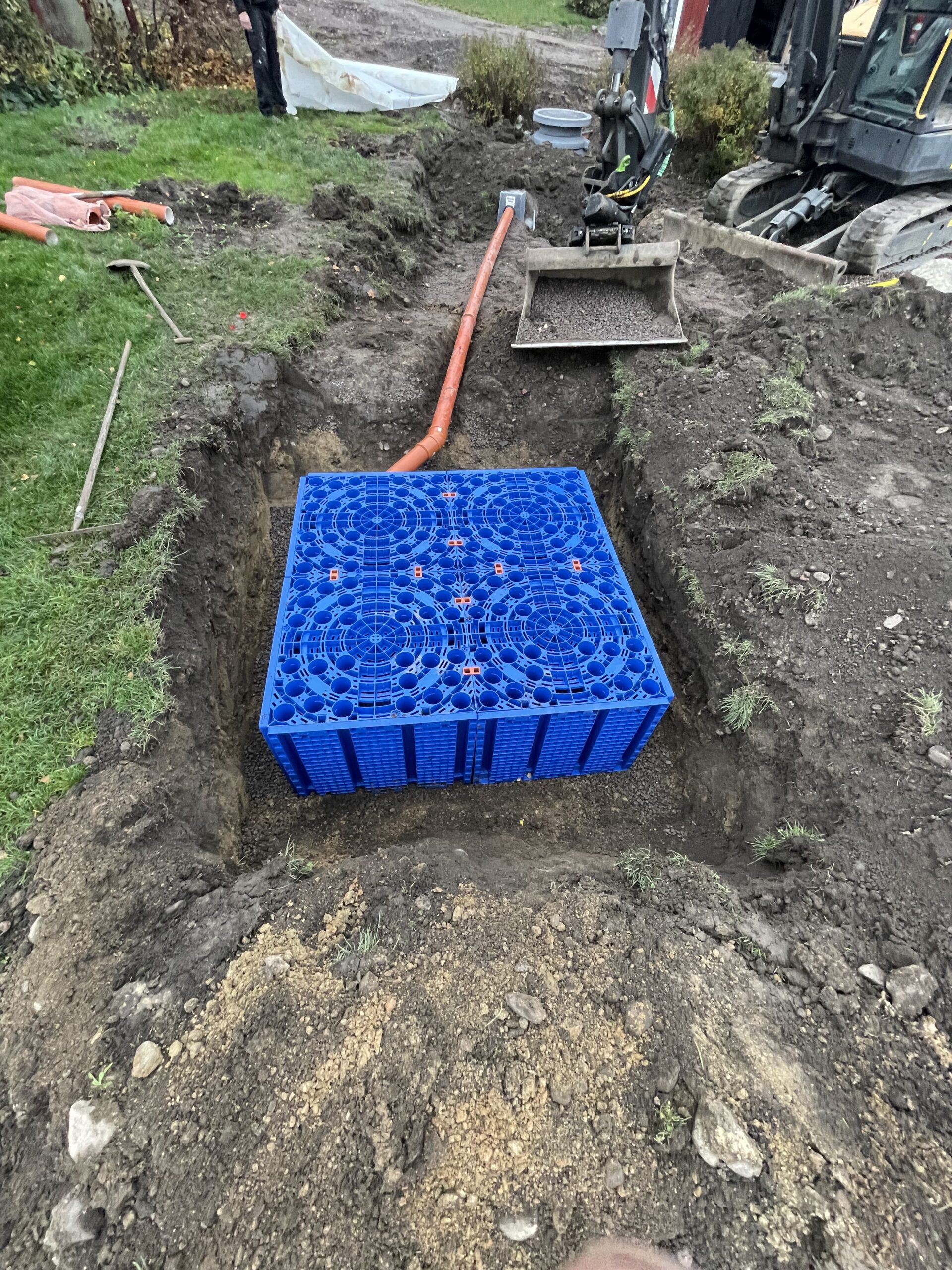 En Fördröjningsmagasin 1600 liter är placerad i ett rektangulärt dike omgivet av jord och gräs, ansluten till ett orange rör, med byggutrustning synlig i bakgrunden.