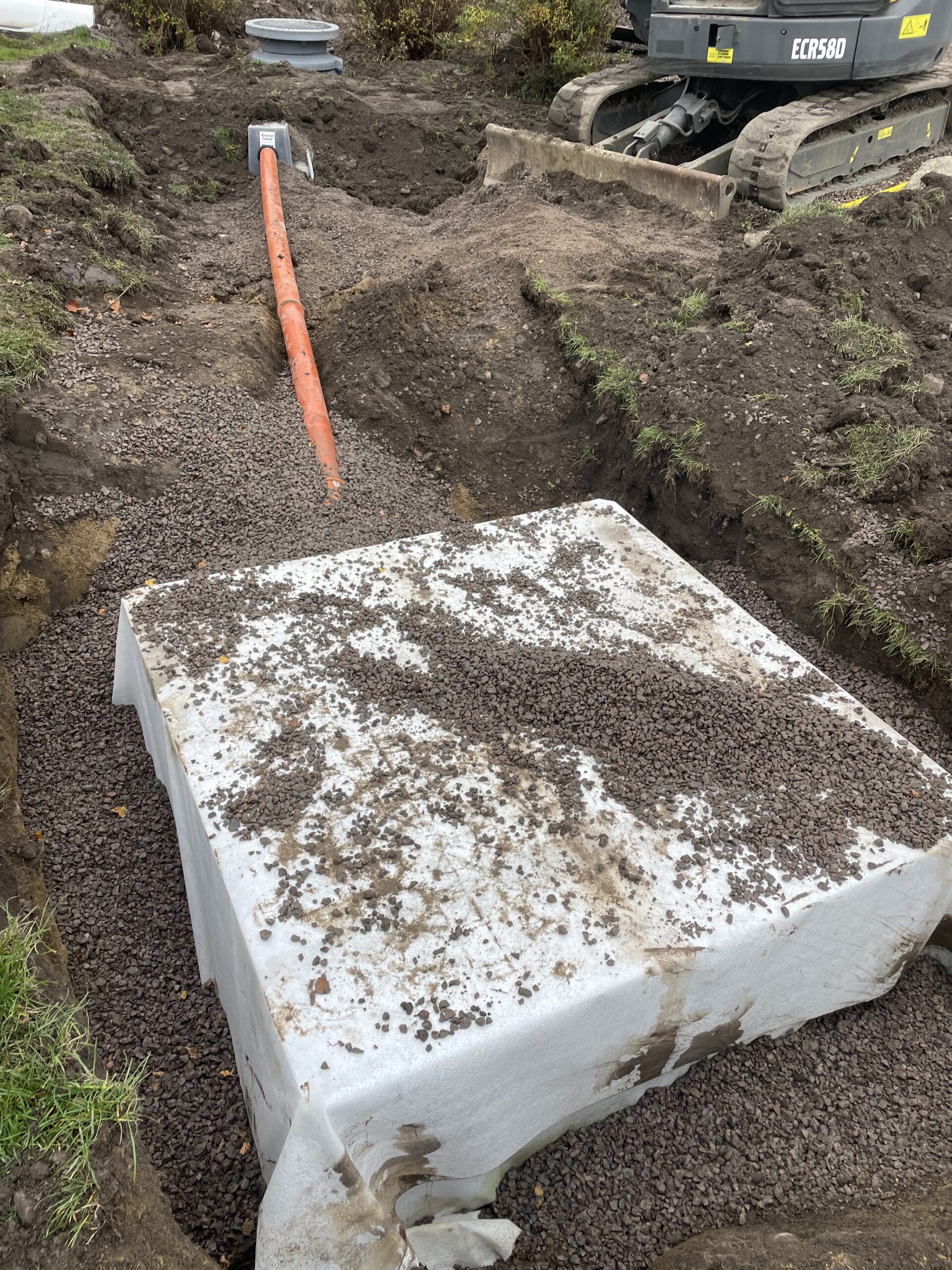 På en byggarbetsplats står en stor vit Fördröjningsmagasin 1600 liters tank delvis nedgrävd i grus, med ett orange rör på. Området är omgivet av jord och smuts och byggutrustning syns i bakgrunden.