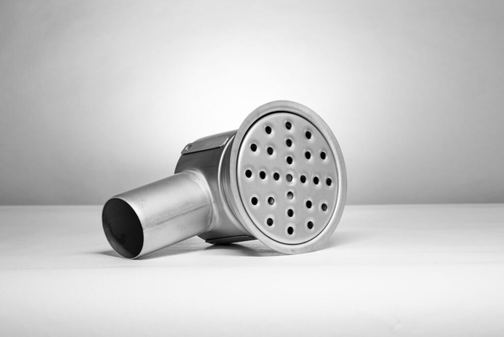 En Rostfria golvbrunnar & rännor med cylindrisk bas och platt, rund perforerad topp ligger i sidled på en vit yta mot en ljusgrå bakgrund.