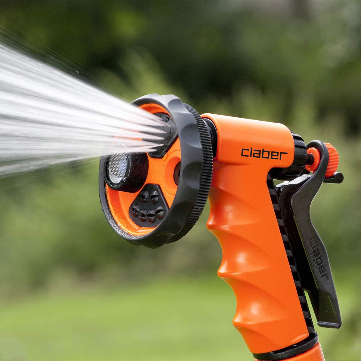 En Claber Ergo-Garden-sprutpistol i orange sprutar vatten mot en suddig grön bakgrund; den har ett svart justerbart munstycke från vilket synliga vattenstrålar strömmar ut.