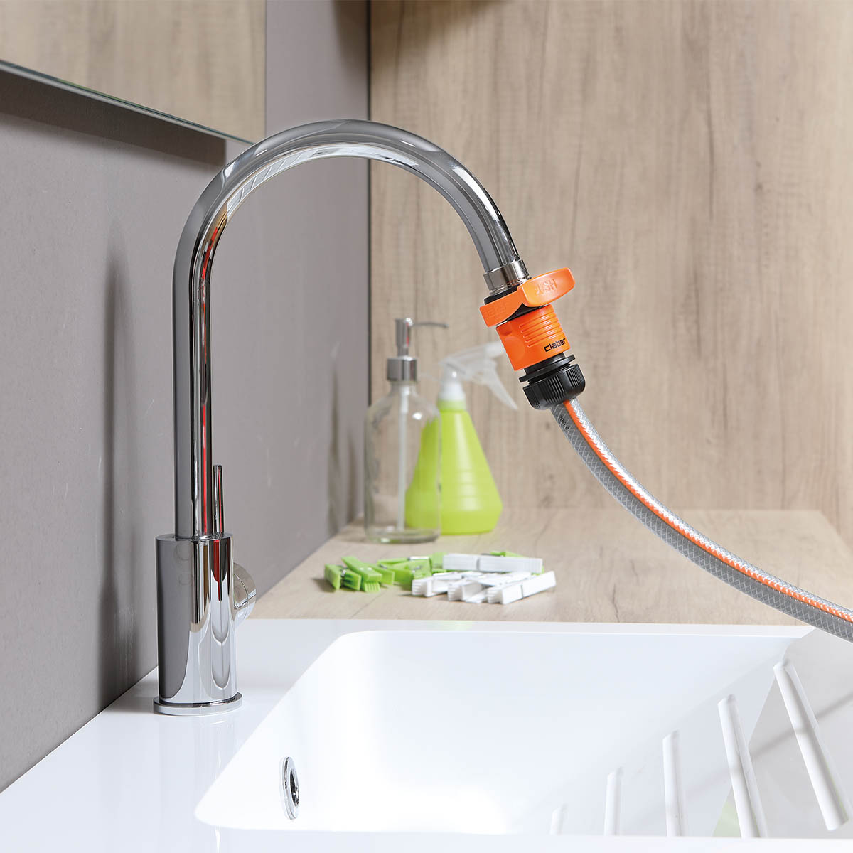 En kromfärgad köksblandare är försedd med en Claber Quick-fit-slangkoppling 3/4″ (orange och svart slangadapter) som är ansluten till en grå trädgårdsslang. Diskbänken är vit, och i bakgrunden syns diskställ och städredskap på träbänkskivan.