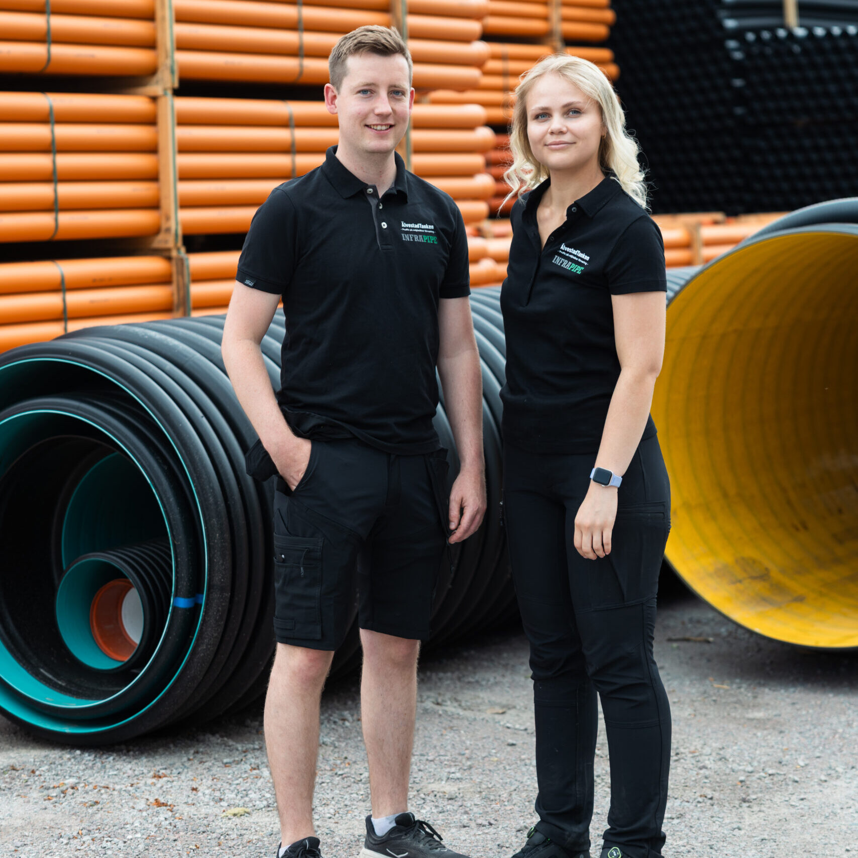 Två personer i svarta kläder står utomhus och ler mot kameran framför stora staplar av PP markrör 110 mm x 6 meter industrirör i orange och svart.