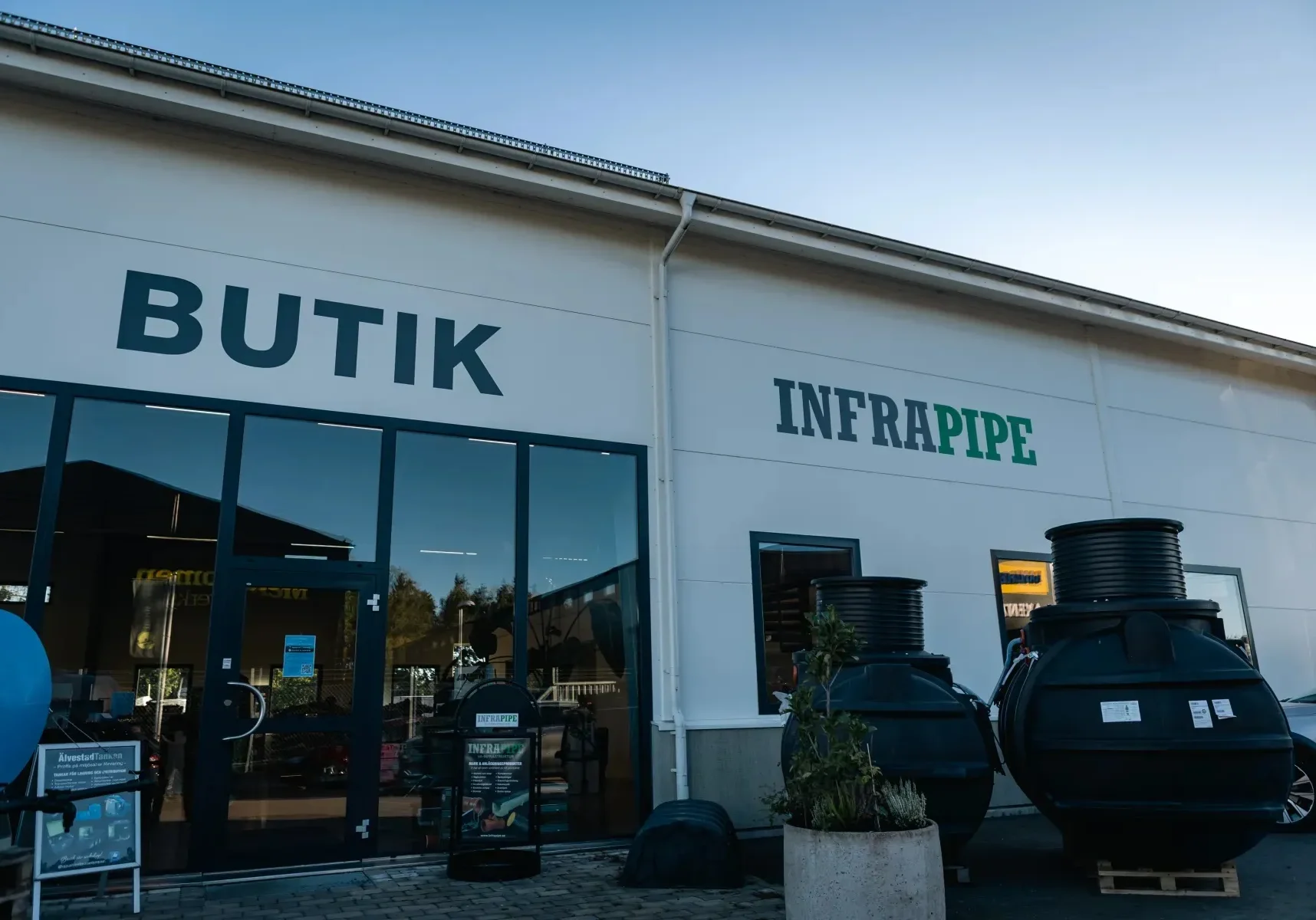 Vy från utsidan av en butik med skyltarna BUTIK och INFRAPIPE. Stora svarta industrirör eller tankar visas utomhus bredvid byggnaden under en klar himmel.