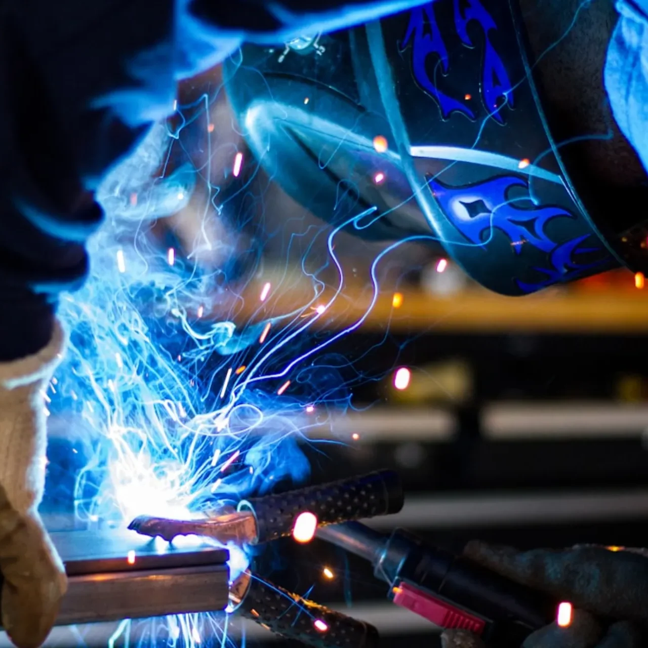 En person iförd skyddshandskar och svetshjälm svetsar metall, och ljusblå gnistor och ljusblixtar lyser upp platsen.